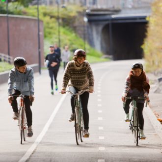 Three smiling people cycling on a wide street, people jogging behind them.