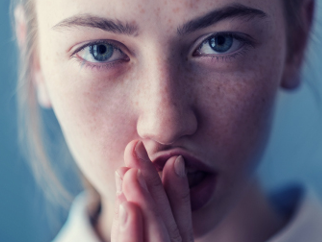 Close-up of a determined-looking person; she’s looking straight to the camera with her hands together in front of her mouth.